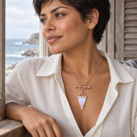 A woman with short dark hair wears a rose gold vermeil necklace and an open white shirt beside a window overlooking the sea.