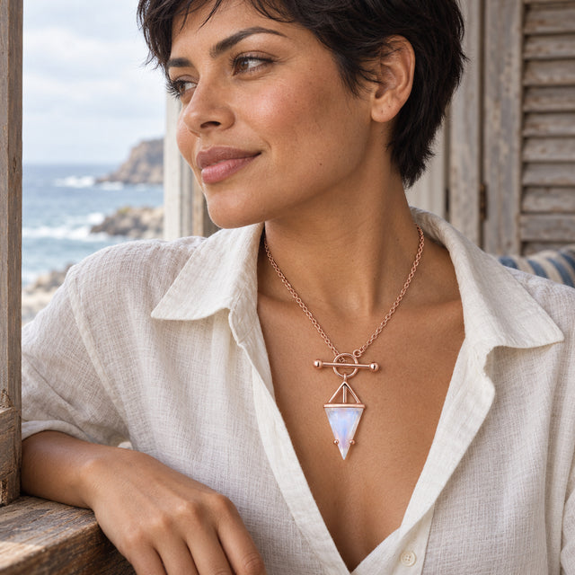 A woman with short dark hair wears a rose gold vermeil necklace and an open white shirt beside a window overlooking the sea.