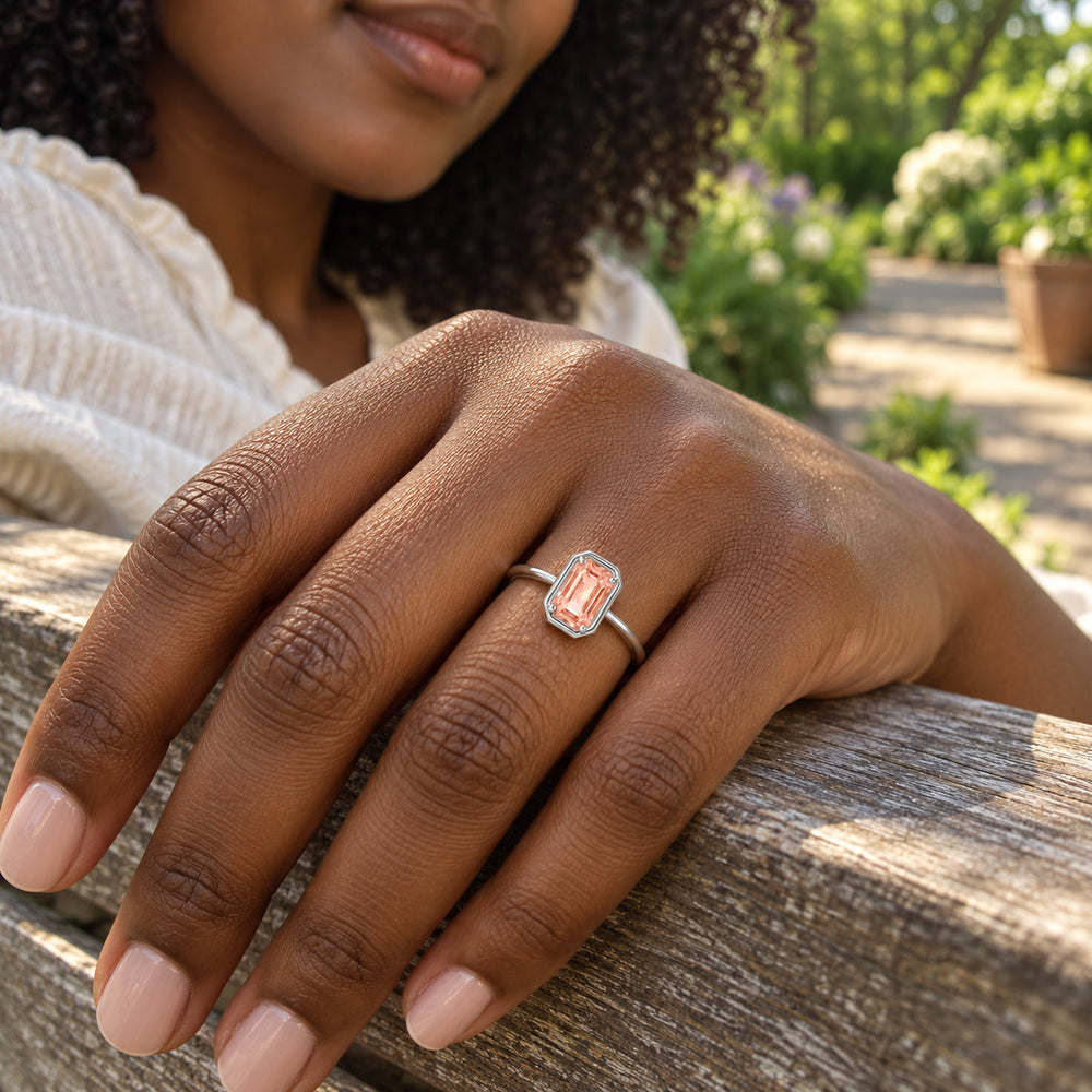 A sterling silver ring with a rectangular peach stone is worn on a hand resting on a wooden garden bench.