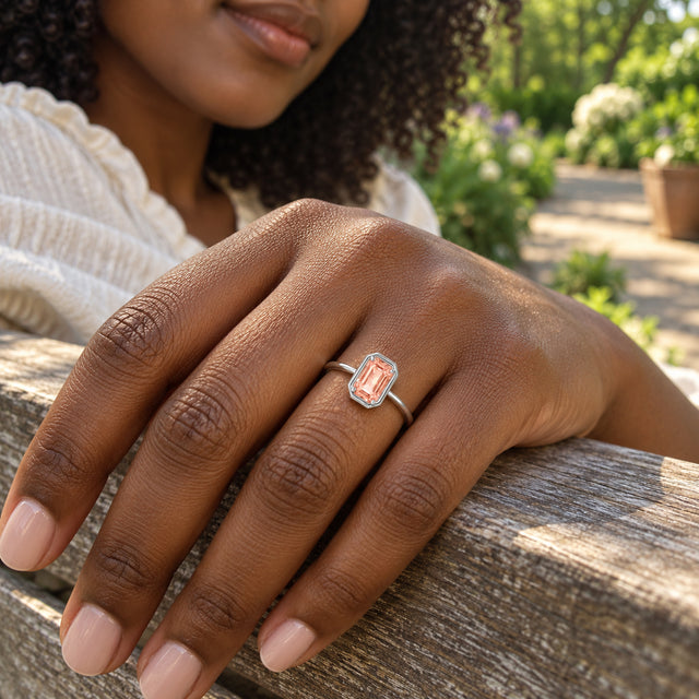 A sterling silver ring with a rectangular peach stone is worn on a hand resting on a wooden garden bench.