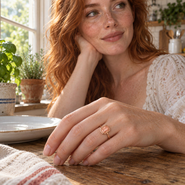 A rose gold vermeil ring with a peach rectangular stone is shown on a hand resting on a wooden kitchen table.
