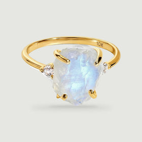 A yellow gold ring holds a rough pale moonstone between two round diamond accents on a light gray surface.