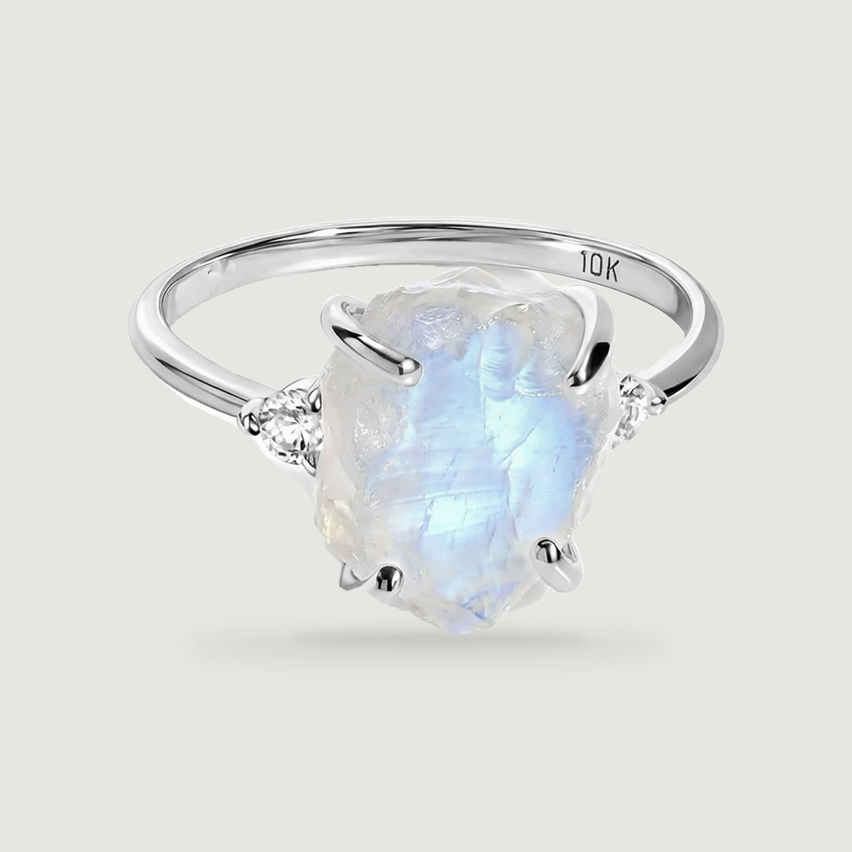 A white gold ring with four rounded prongs frames a blue-flashing rough moonstone and two side diamonds.