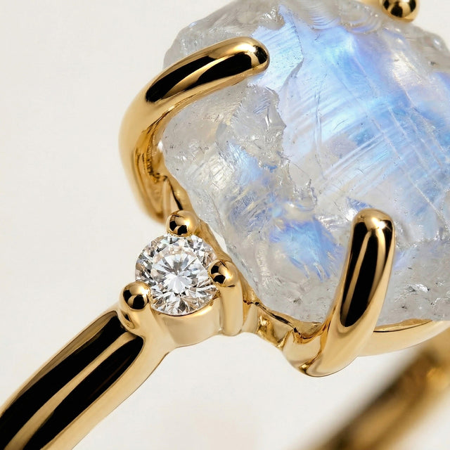 A close view shows a yellow gold prong, a round diamond, and the textured blue-flashing surface of the moonstone.