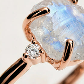 Polished rose gold prongs and a round diamond sit against the clear ridges and blue reflections of the rough moonstone.