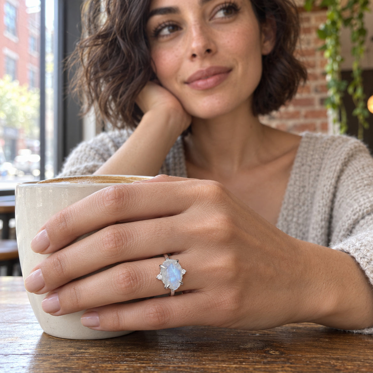 A woman with a wavy brown bob wears the white gold moonstone ring while holding a ceramic cup at a cafe table.