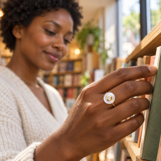 A yellow gold vermeil moonstone ring is worn on a hand reaching toward books on a wooden shelf.