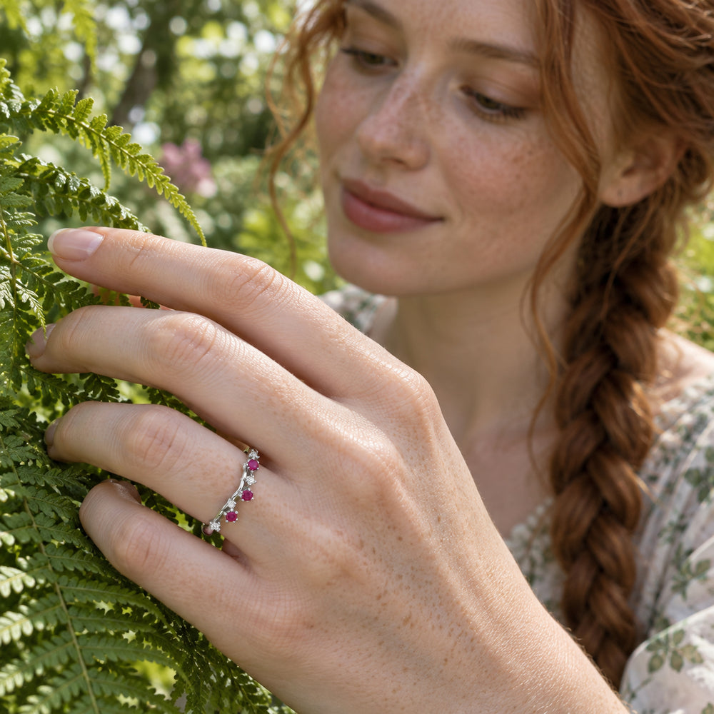 A silver ring with pink-red and clear stones is worn on a freckled hand touching a fern.