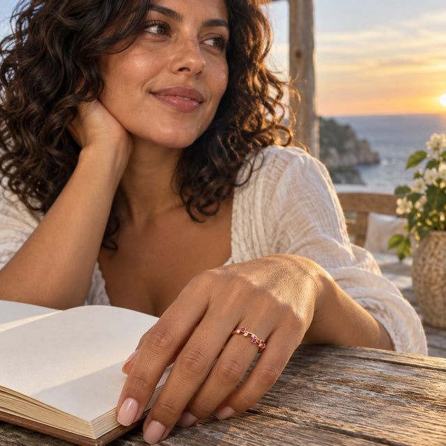 A rose gold vermeil ring with pink-red stones is shown on a hand resting beside an open book on a wooden table.