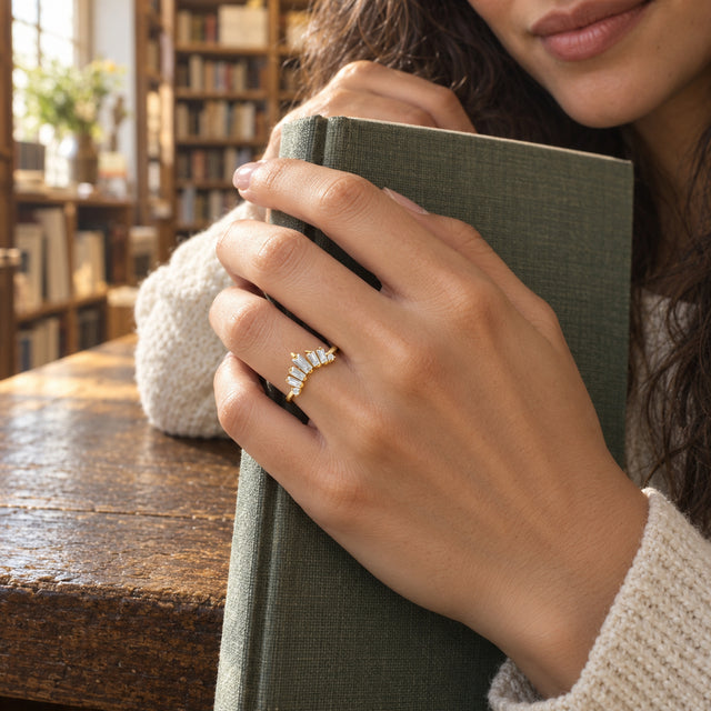 A yellow gold vermeil ring is worn on a hand holding a green clothbound book in a sunlit bookshop.