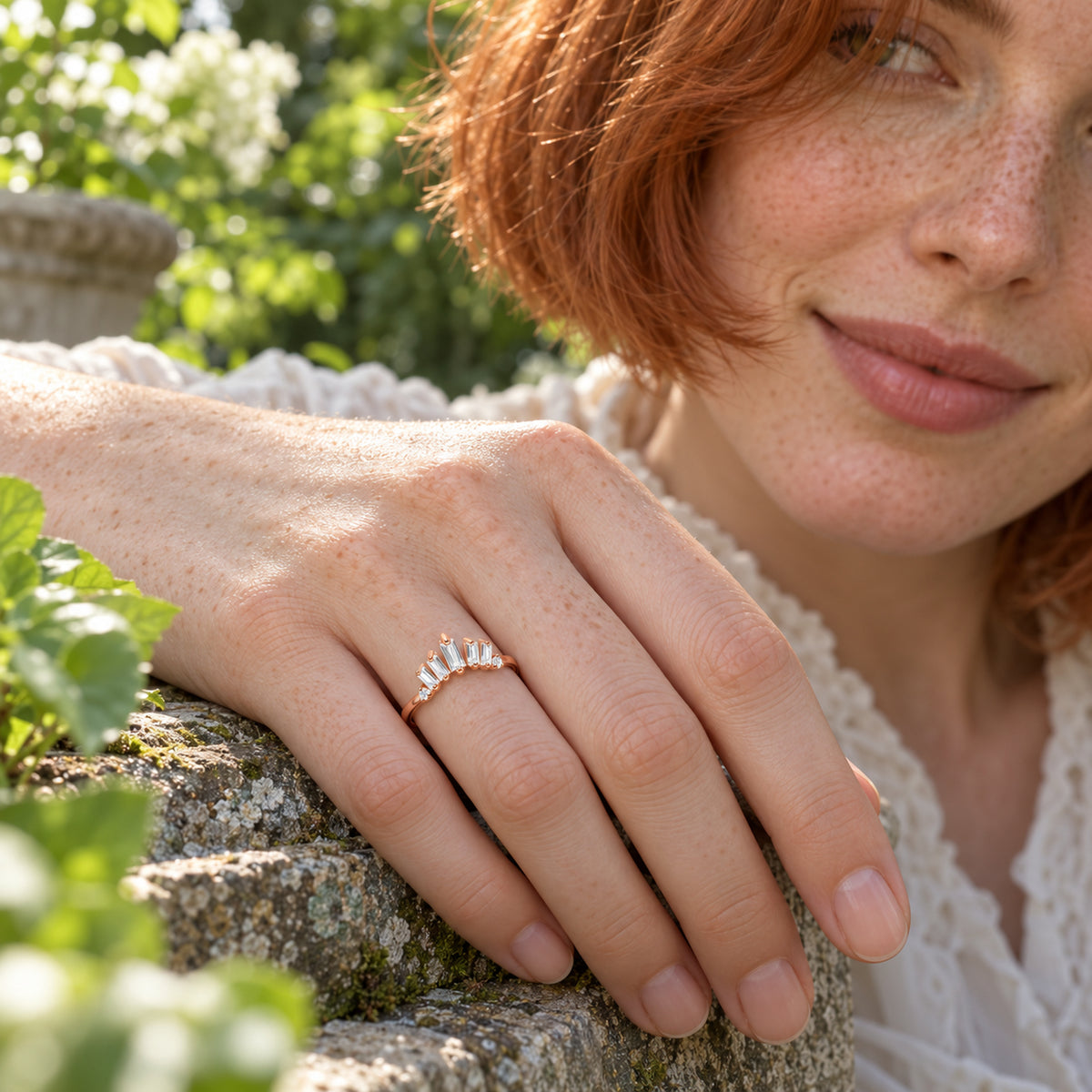 A rose gold vermeil band is worn on a freckled hand resting on a weathered stone ledge among green leaves.
