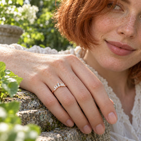 A rose gold vermeil band is worn on a freckled hand resting on a weathered stone ledge among green leaves.