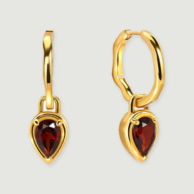 Two polished yellow gold hoops have pear-shaped deep red garnet drops on a light gray background.