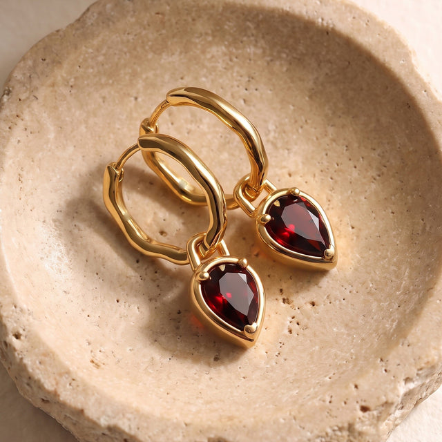 Two yellow gold garnet hoop earrings rest diagonally on a textured pale stone dish.