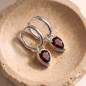 Silver-tone hoops with red garnet pendants lie together on a warm beige stone surface.