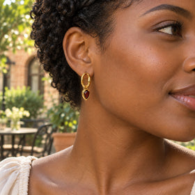A woman with short curls wears a yellow gold garnet hoop earring in a sunlit courtyard.