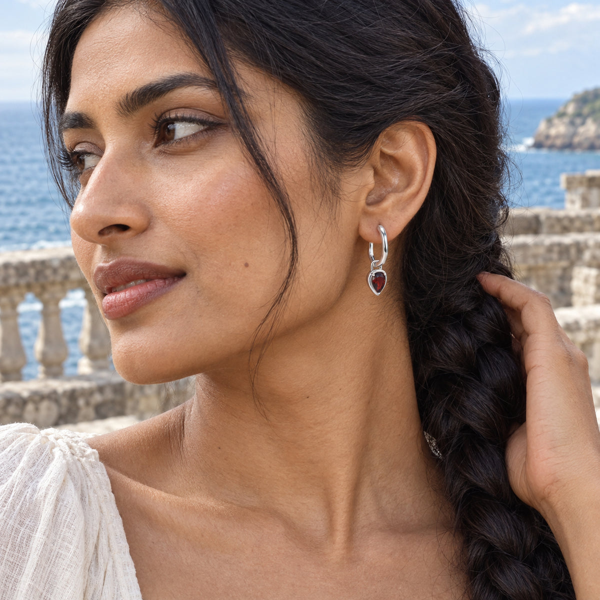 A woman with a dark braid wears a silver-tone garnet hoop beside a stone terrace overlooking the sea.