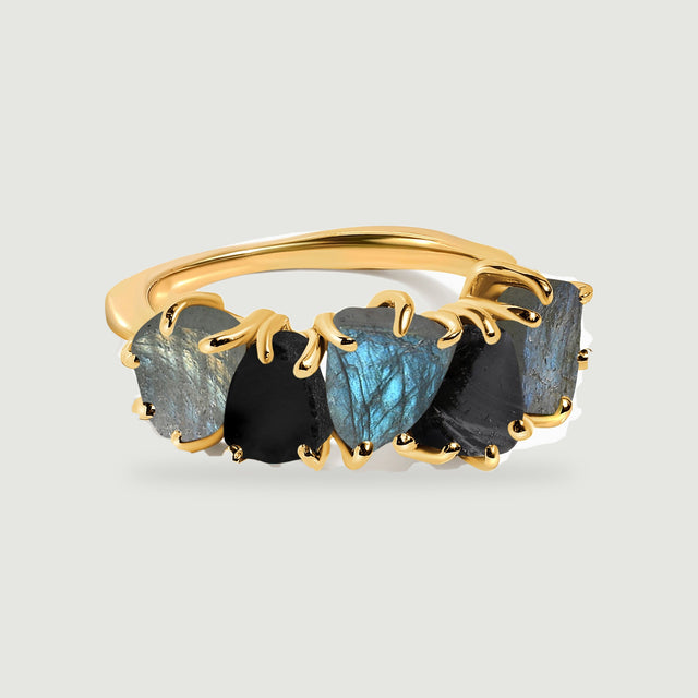 A yellow gold-tone ring with blue-gray and black raw stones rests centrally on a pale gray background.