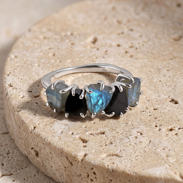The five-stone silver-tone ring rests on a textured pale stone block with its curved band visible behind the gems.