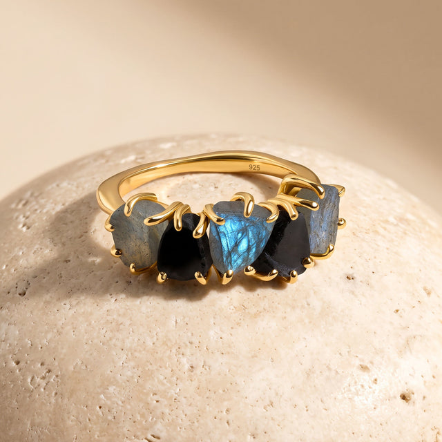 A yellow gold-tone band curves behind alternating gray-blue labradorite and black obsidian on a light stone surface.