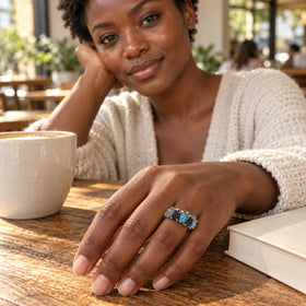 A silver-tone five-stone ring is worn on a hand resting on a cafe table beside a ceramic cup and a closed book.