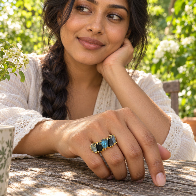 A yellow gold-tone ring with blue-gray and black raw stones is worn on a hand resting on a wooden garden table beside white flowers.