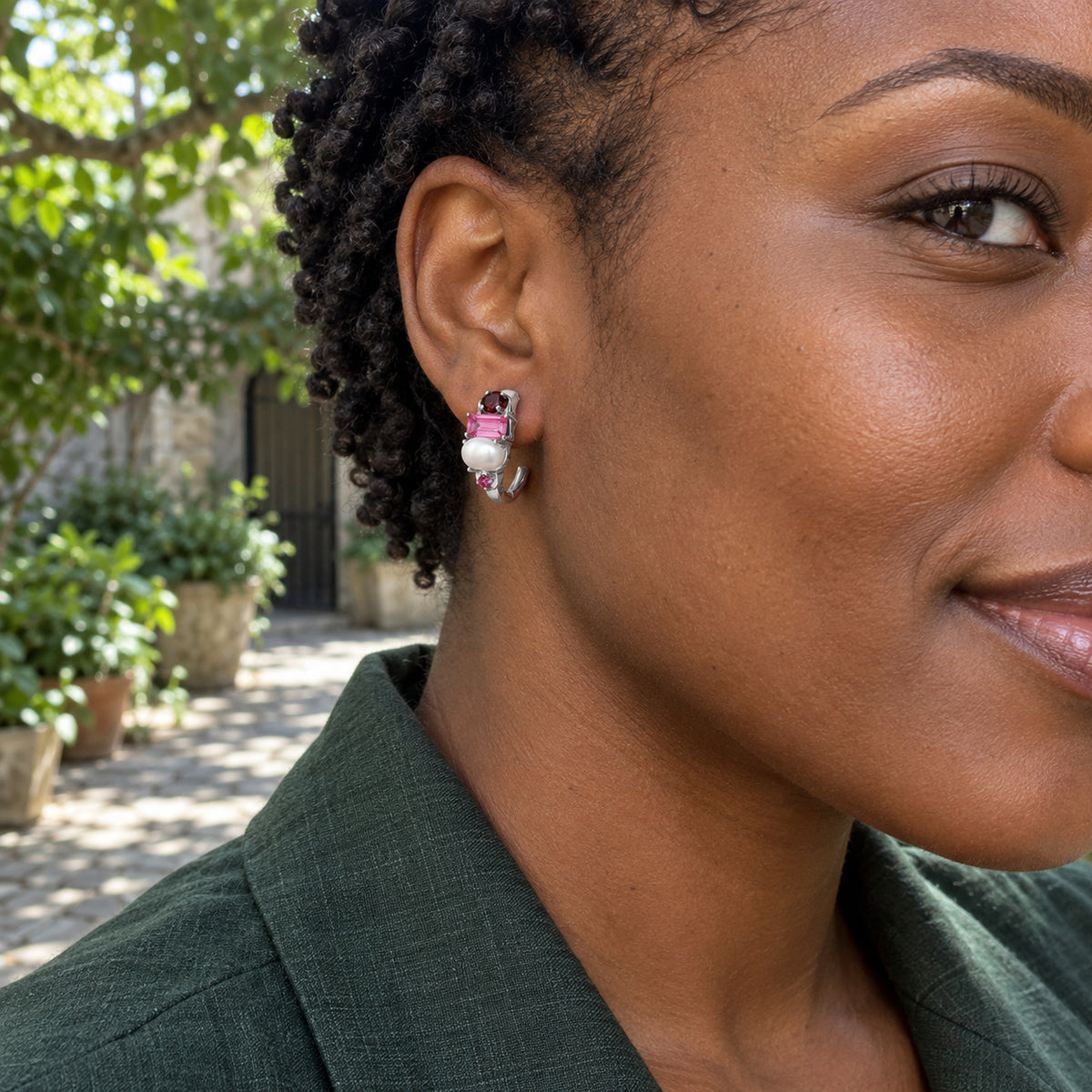 A woman with short twists wears a silver-tone pearl and garnet hoop in a leafy courtyard.