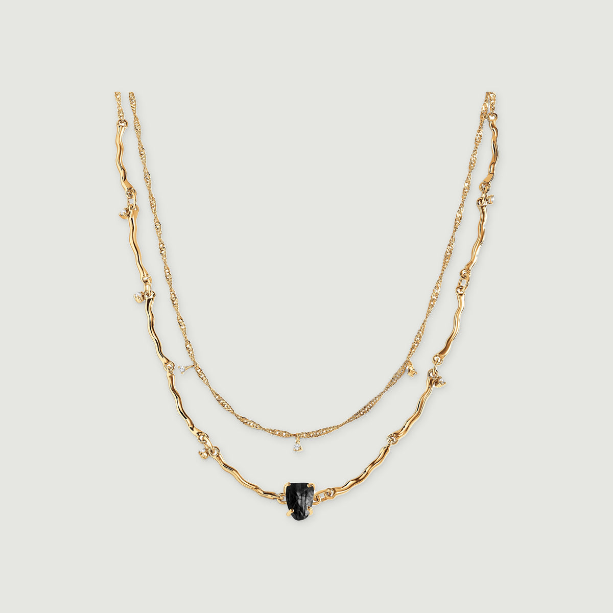 A yellow gold double-chain necklace with a raw black stone and small clear stone charms rests on a light grey surface.