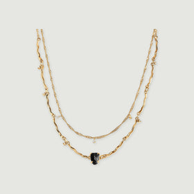 A yellow gold double-chain necklace with a raw black stone and small clear stone charms rests on a light grey surface.