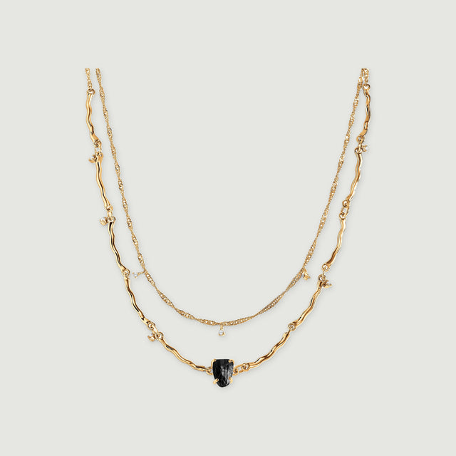 A yellow gold double-chain necklace with a raw black stone and small clear stone charms rests on a light grey surface.