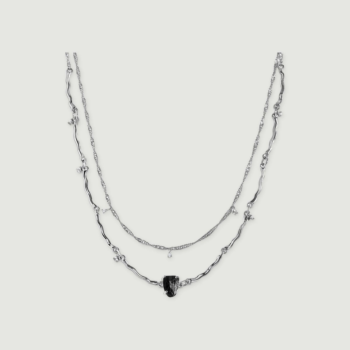 A silver double-chain necklace with curved links, a raw black stone and tiny clear stone drops is centered on light grey.