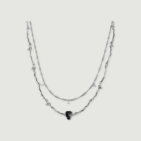 A silver double-chain necklace with curved links, a raw black stone and tiny clear stone drops is centered on light grey.