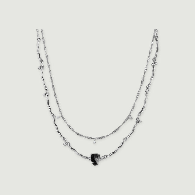 A silver double-chain necklace with curved links, a raw black stone and tiny clear stone drops is centered on light grey.