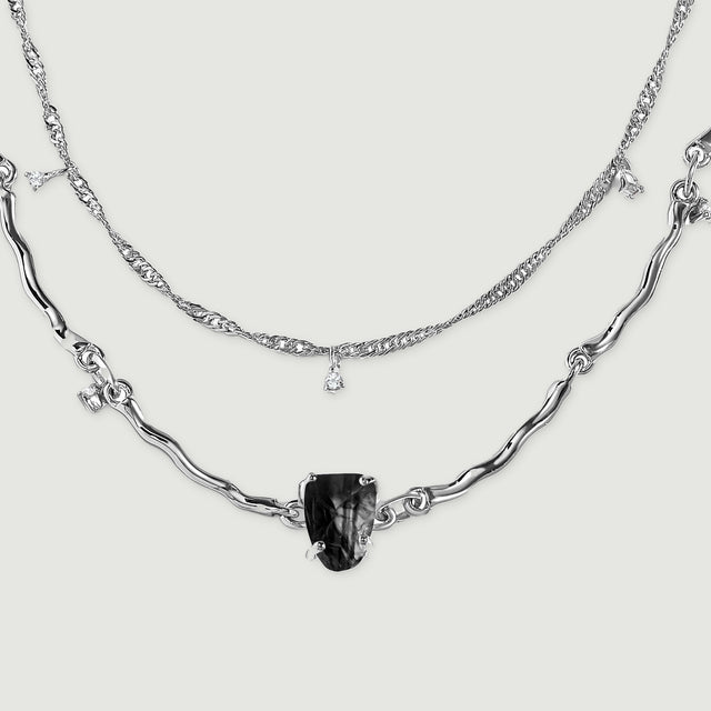 Two silver chain arcs lie flat, showing textured fine links above a sculpted strand with a black center stone.