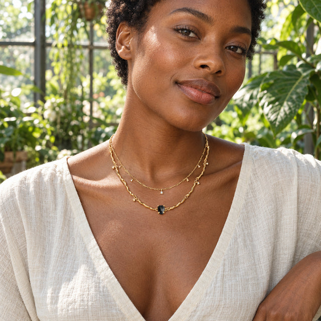 A woman in an ivory linen blouse wears the yellow gold two-strand black obsidian necklace in a leafy greenhouse.