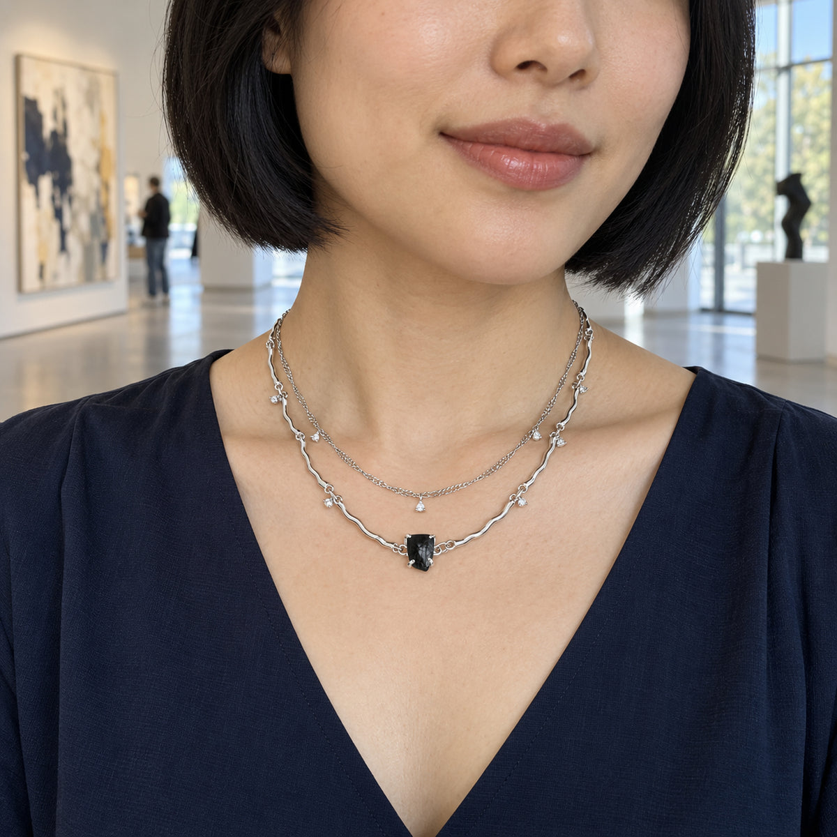 A silver two-strand necklace with a black center stone and tiny clear charms rests above a navy V-neck blouse in an art gallery.