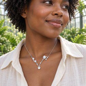 A woman in a cream linen shirt wears a silver necklace with four pale blue stones inside a greenhouse.