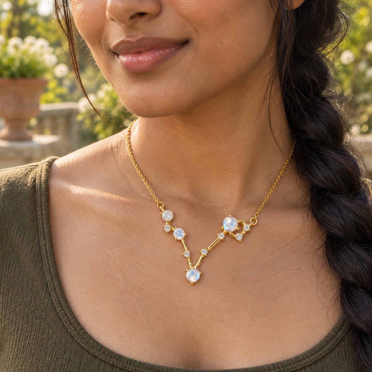 A yellow gold necklace with an angular four-stone pendant rests above an olive scoop neckline on a garden terrace.