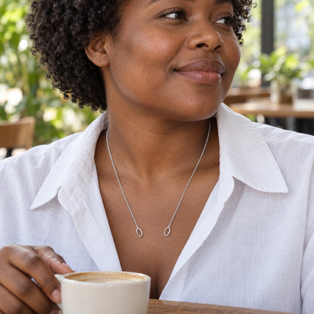 A woman in a white shirt wears a delicate silver chain with two oval ends while holding a coffee cup in a leafy cafe.