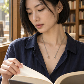 A woman with a short black bob wears the silver chain above a navy blouse while turning a book page beside wooden shelves.