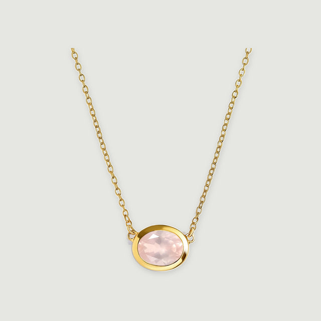 A yellow gold colored bezel surrounds a pale pink oval stone beneath two lengths of matching link chain on light gray.