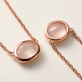 Rose gold colored oval bezels and fine chain connections frame two faceted pink stones in a close view on a cream surface.