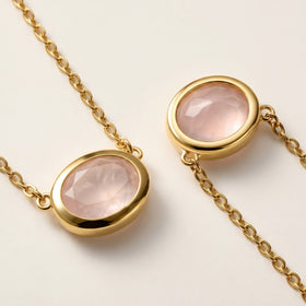A close view shows the yellow gold colored settings and translucent pink facets of two oval pendants resting on a cream tabletop.