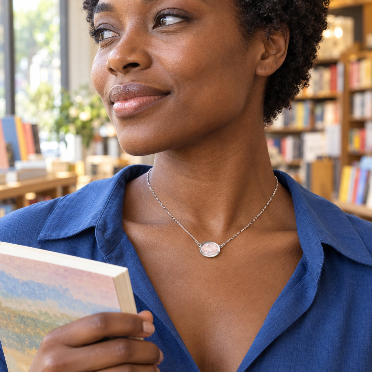 A woman with short curly hair wears a white metal pink oval pendant over a blue blouse while holding a book beside a window.