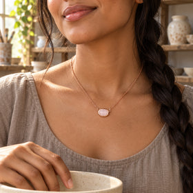 A woman with a long dark braid wears a rose gold colored pink oval necklace over a taupe linen top while resting her hand on a ceramic bowl.