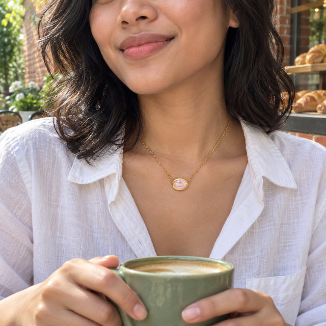 A woman with wavy black hair wears a yellow gold colored oval pink pendant with a white shirt while holding a green cup outside a brick bakery.