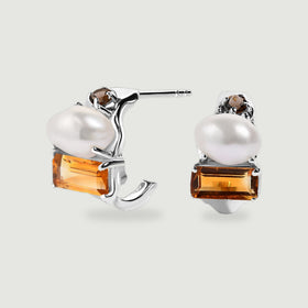 A pair of silver-tone open hoop earrings displays white pearls, rectangular amber-colored citrine and small round brown stones on a light gray surface.