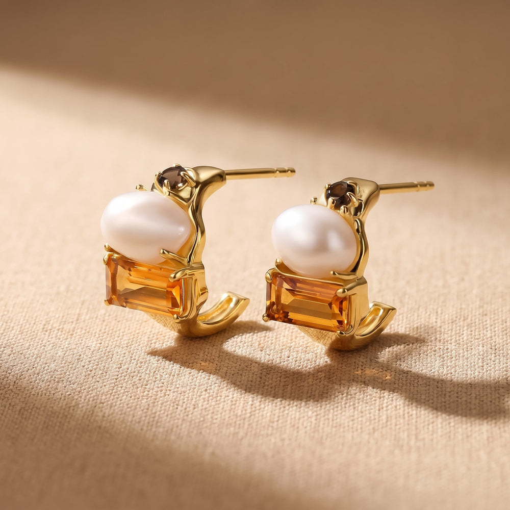 Two yellow gold earrings rest on beige woven fabric with their pearls and rectangular orange stones facing forward.