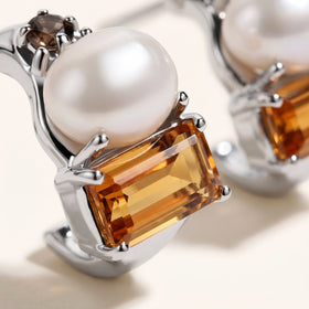 Polished silver-tone prongs frame an orange faceted citrine below a white pearl and a small brown gemstone.