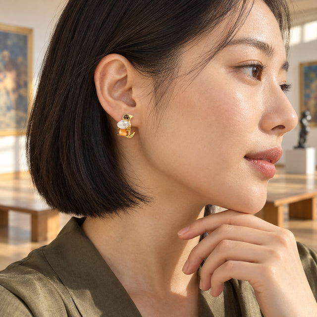 A woman with a short black bob wears a yellow gold pearl and citrine earring against a softly blurred gallery interior.
